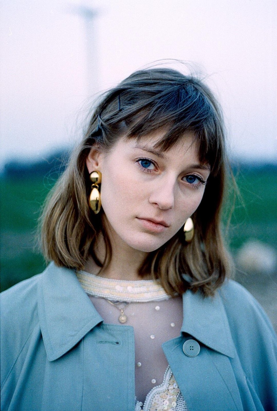 A woman with blue eyes and gold earrings wears a blue coat and a sheer top with sequins.