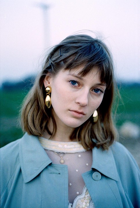 A woman with blue eyes and gold earrings wears a blue coat and a sheer top with sequins.