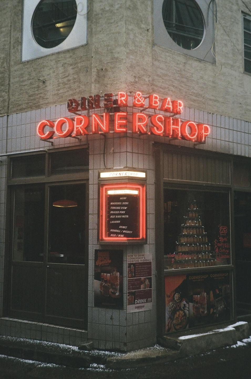 A neon sign for a diner and bar called Cornershop illuminates a building at night.