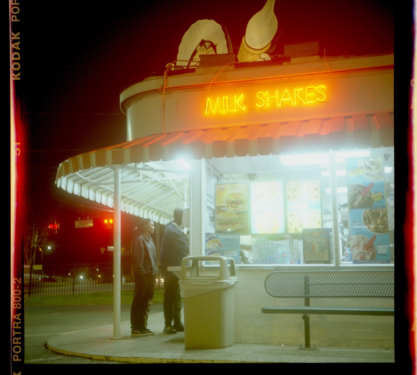 Two people stand outside a brightly lit Dairy Queen at night, with a neon "MILK SHAKES" sign glowing above the service window.