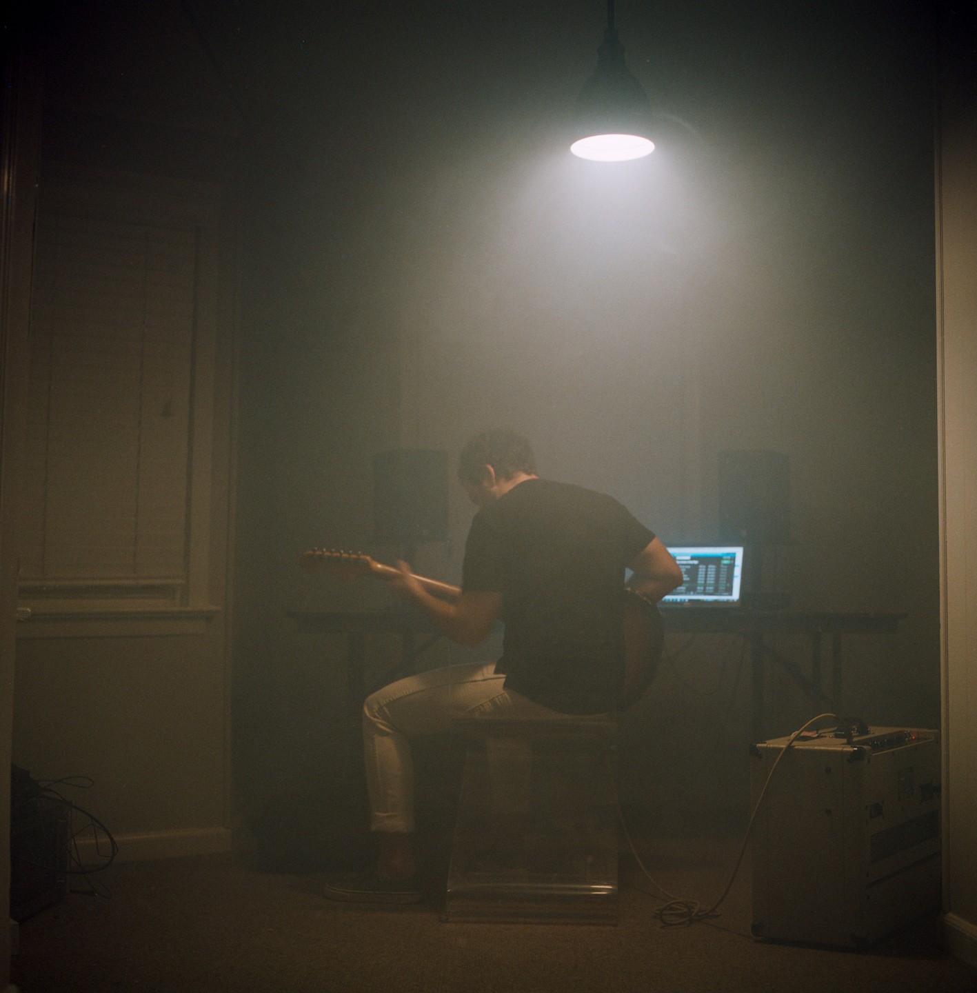 A man sits on a stool playing an electric guitar in a dimly lit room with a laptop and speakers on a table.
