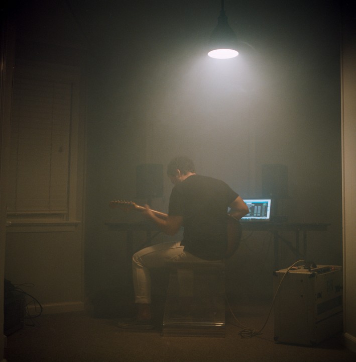 A man sits on a stool playing an electric guitar in a dimly lit room with a laptop and speakers on a table.