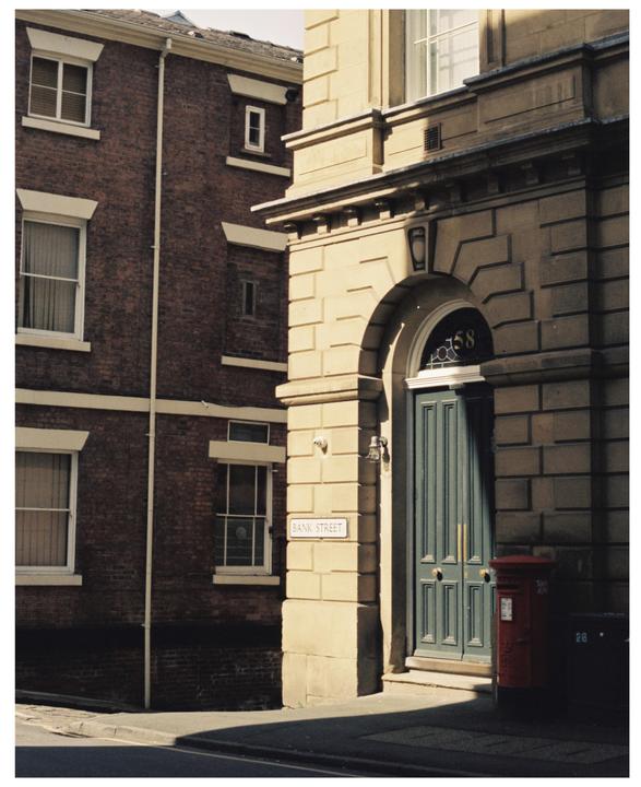 A red mailbox sits next to a green door on a sunlit street corner with a brick building in the background.