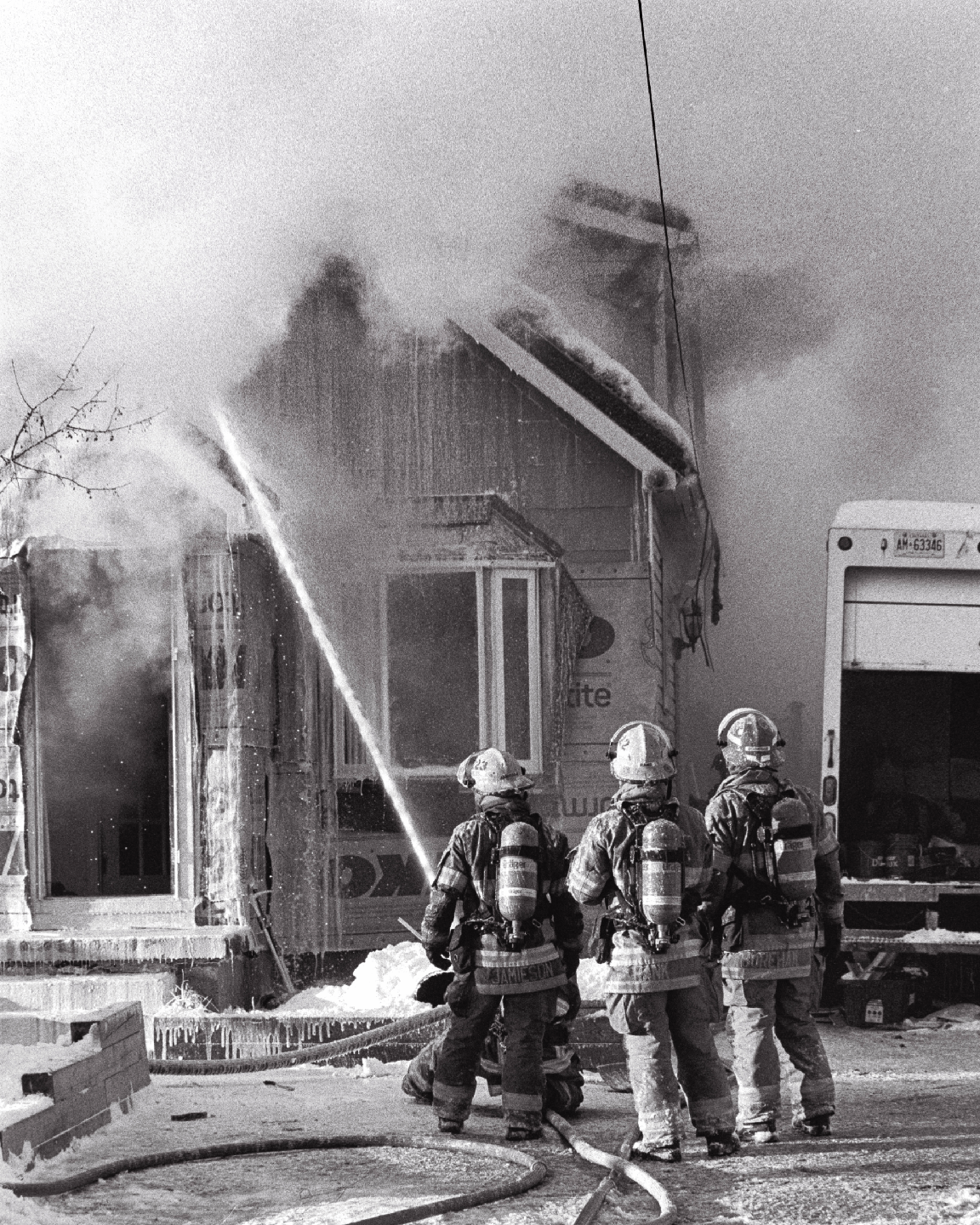 Three firefighters spray water on a burning house covered in snow and ice.