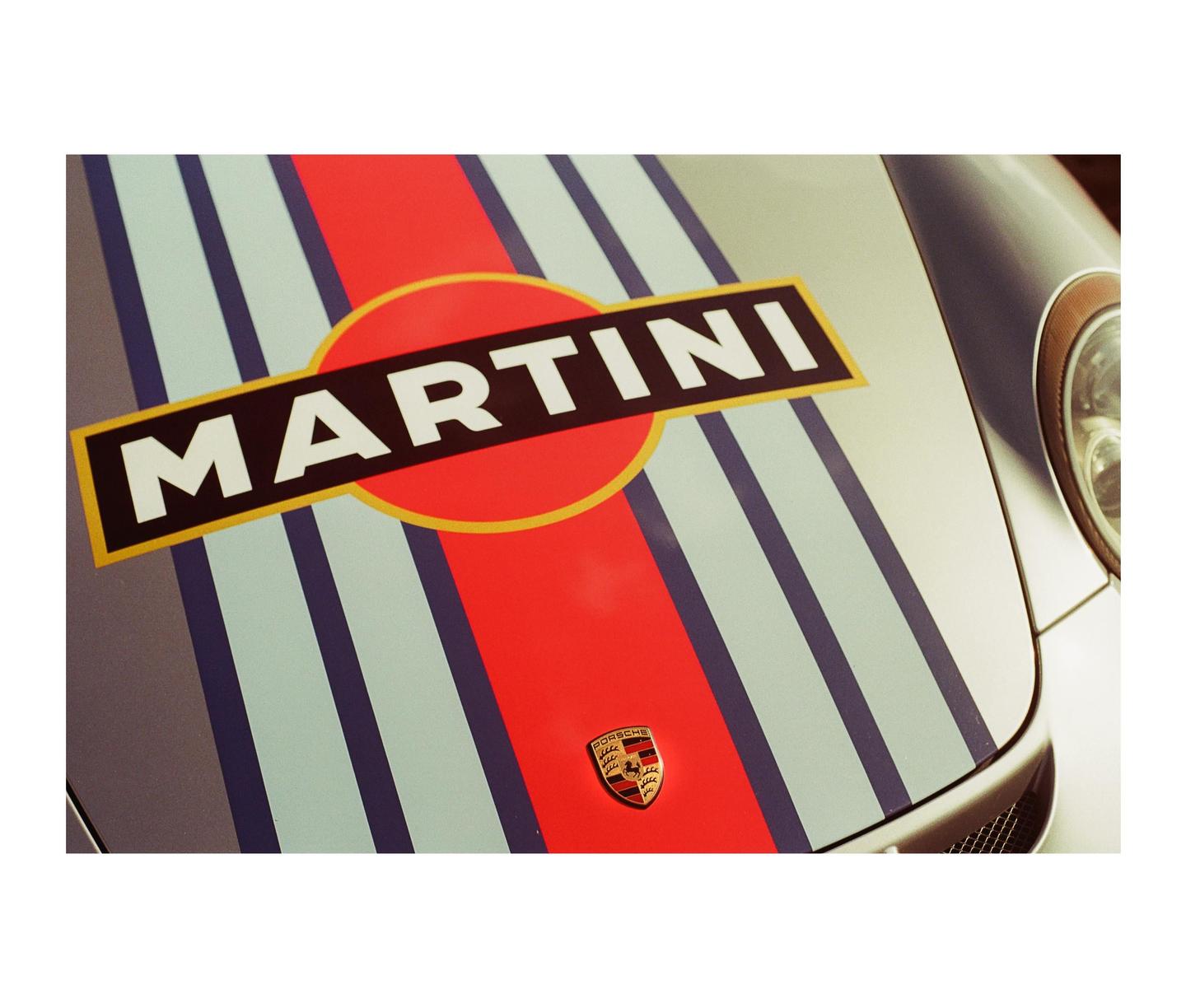 The front of a car is decorated with a Martini racing stripe and a Porsche emblem.