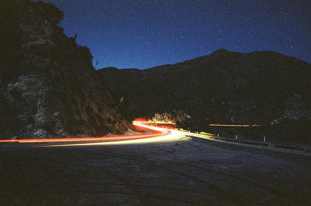 A winding mountain road at night is illuminated by the light trails of passing vehicles under a starry sky.