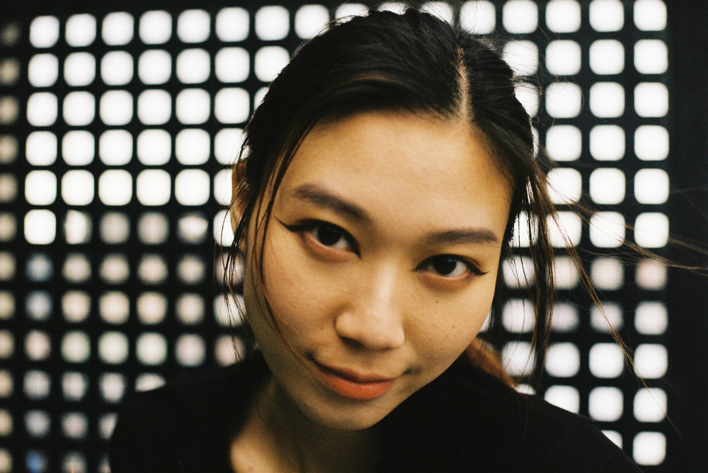 A woman with dark hair and winged eyeliner looks directly at the viewer against a background of illuminated squares.