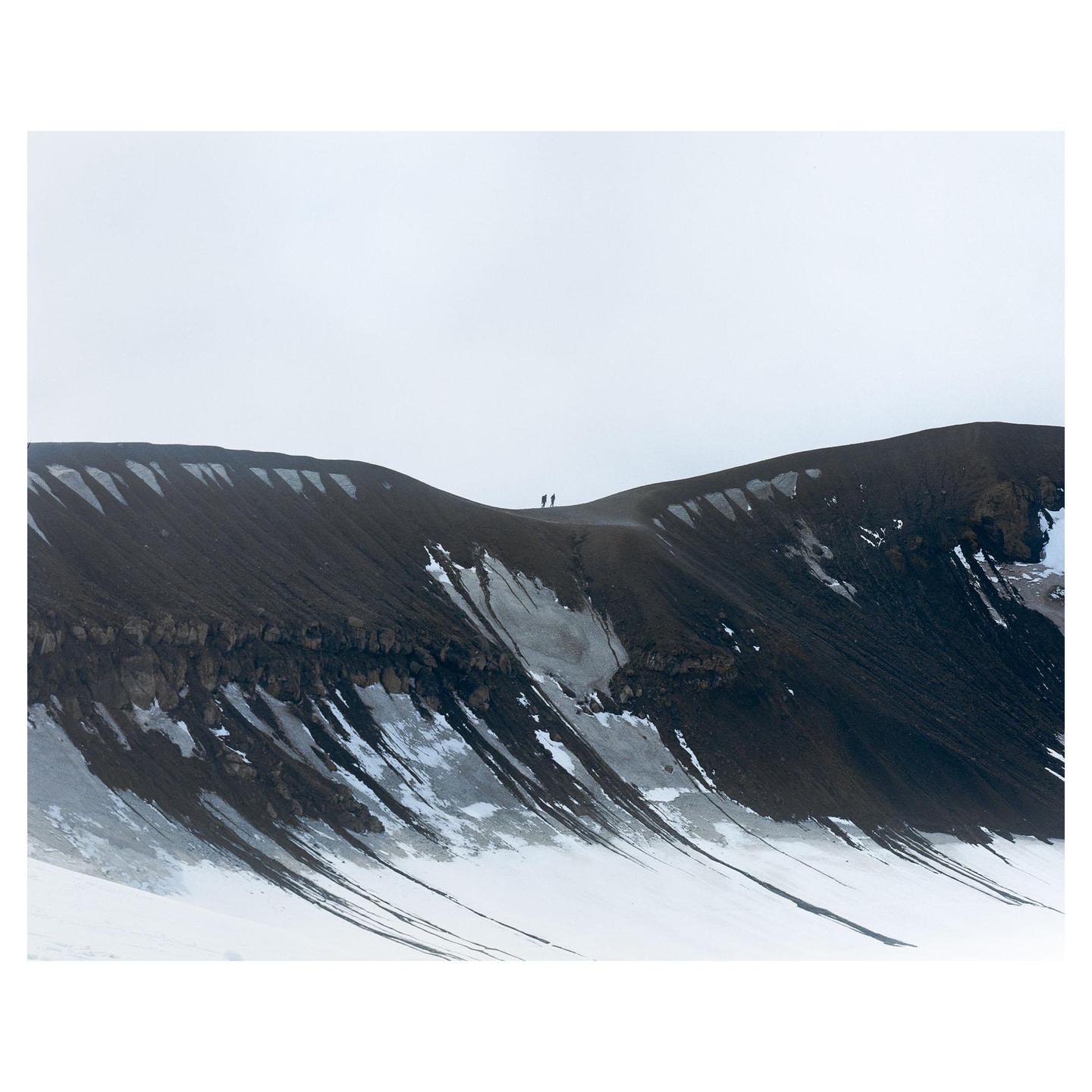Two people stand on a dark, snow-dusted ridge under a pale sky.
