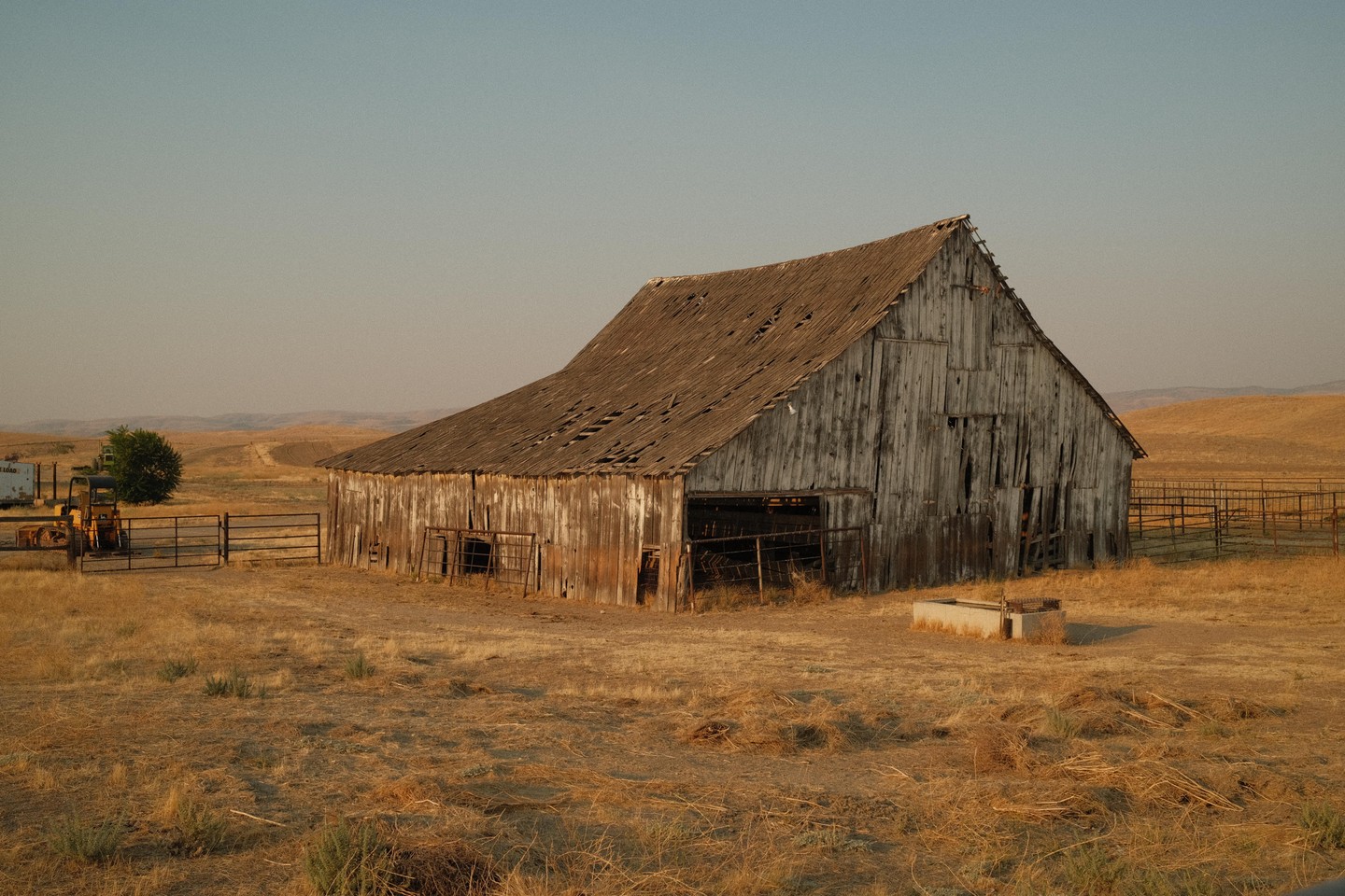 An old wooden barn with a damaged roof stands in a dry, grassy field under a clear sky.