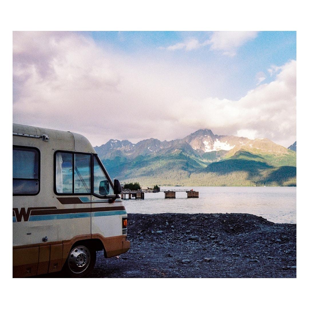 A motorhome is parked on a rocky shore with mountains and a body of water in the background.