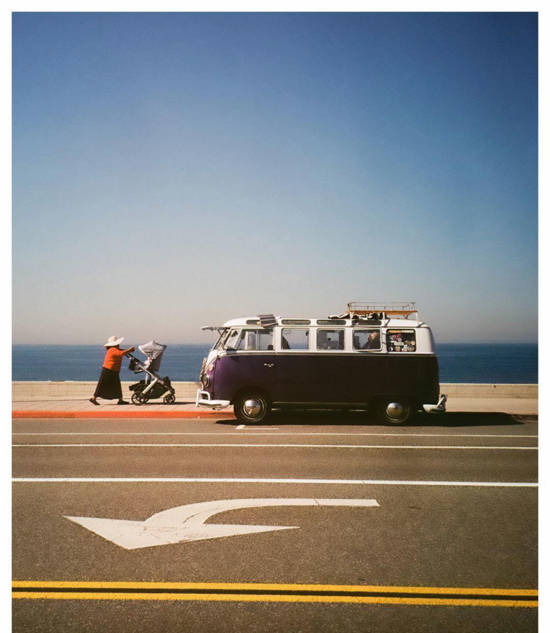 A woman pushes a stroller past a purple and white van parked next to a road with an ocean in the background.