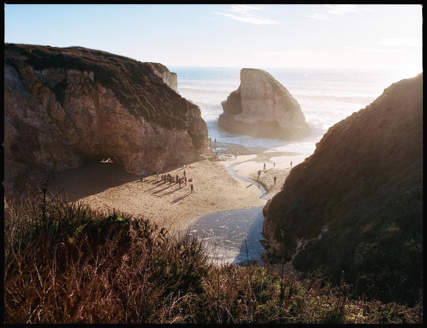 A group of people are gathered on a sandy beach between two large cliffs, with the ocean and a sea stack in the background.