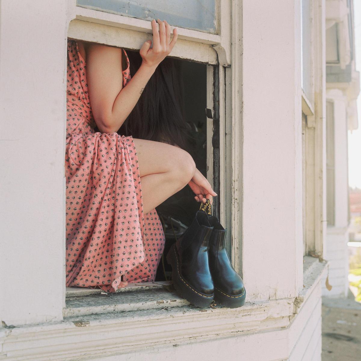 A woman in a pink dress sits in a window frame, holding a pair of black boots.