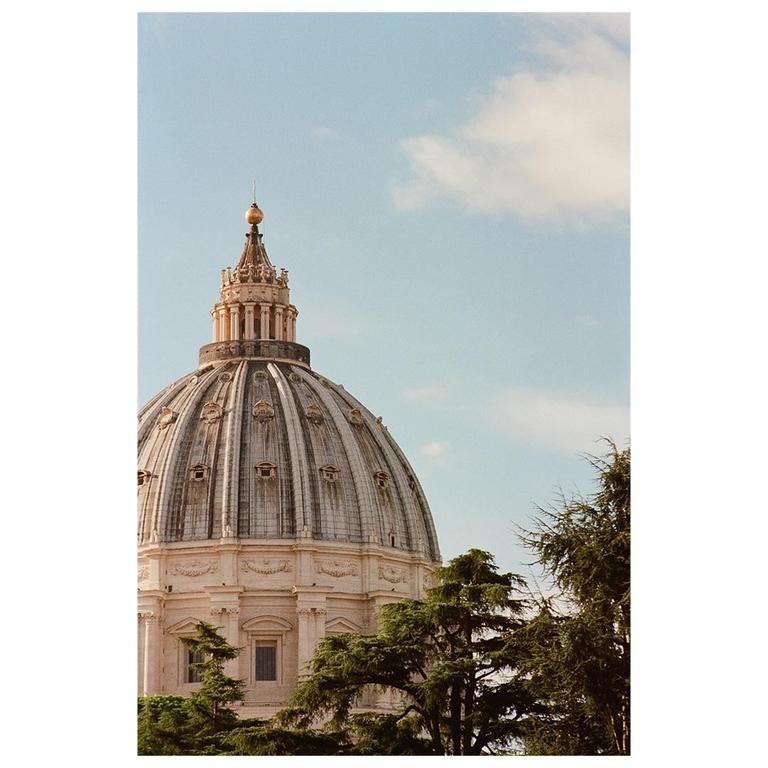The dome of St. Peter's Basilica rises above green trees against a blue sky.