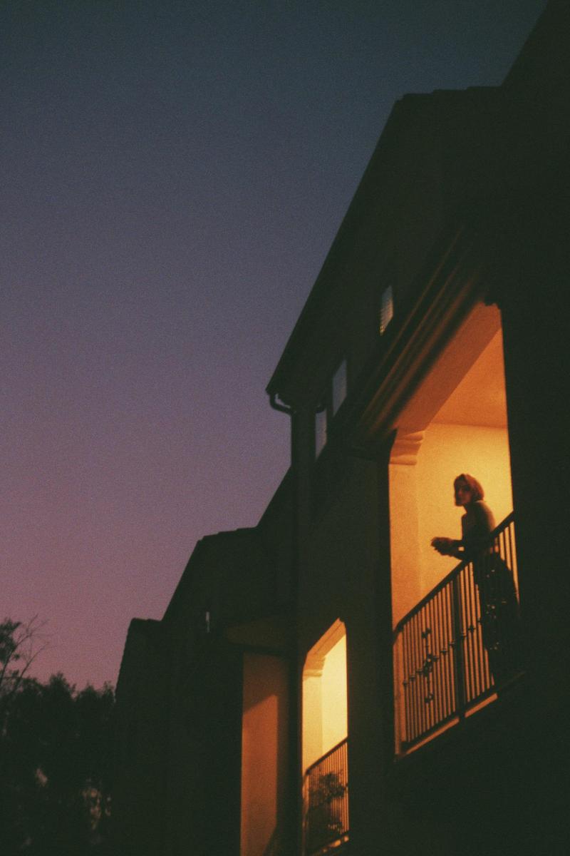 A woman stands on a balcony illuminated by warm light at dusk.