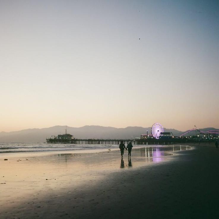 Two people walk on a beach toward a pier with a Ferris wheel and mountains in the background at dusk.