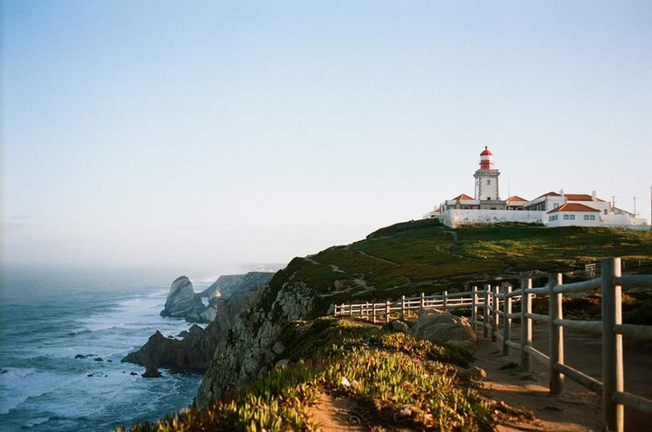 A lighthouse with a red top stands on a grassy cliff overlooking the ocean with a wooden fence in the foreground.