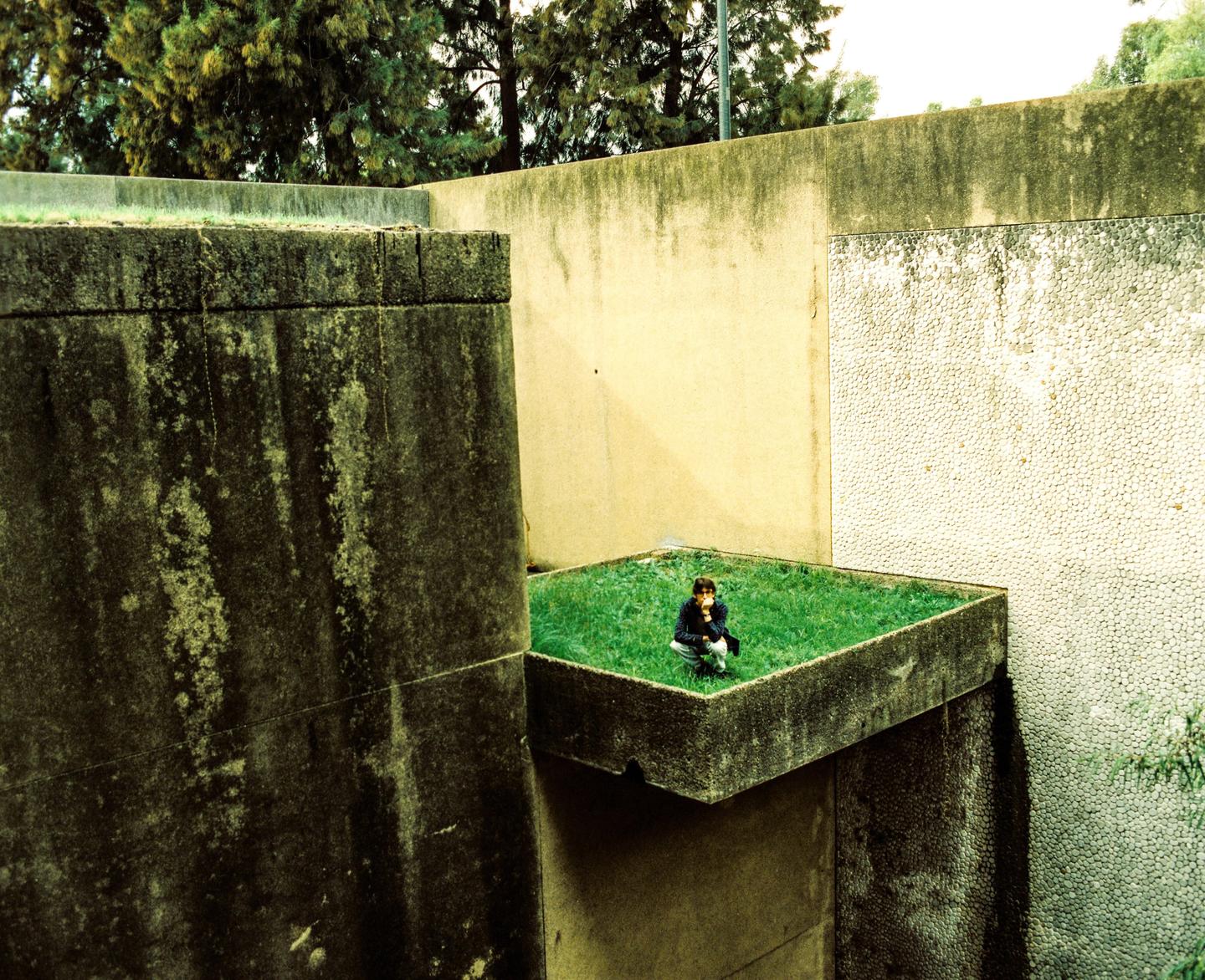 A person crouches on a small patch of grass surrounded by concrete walls.