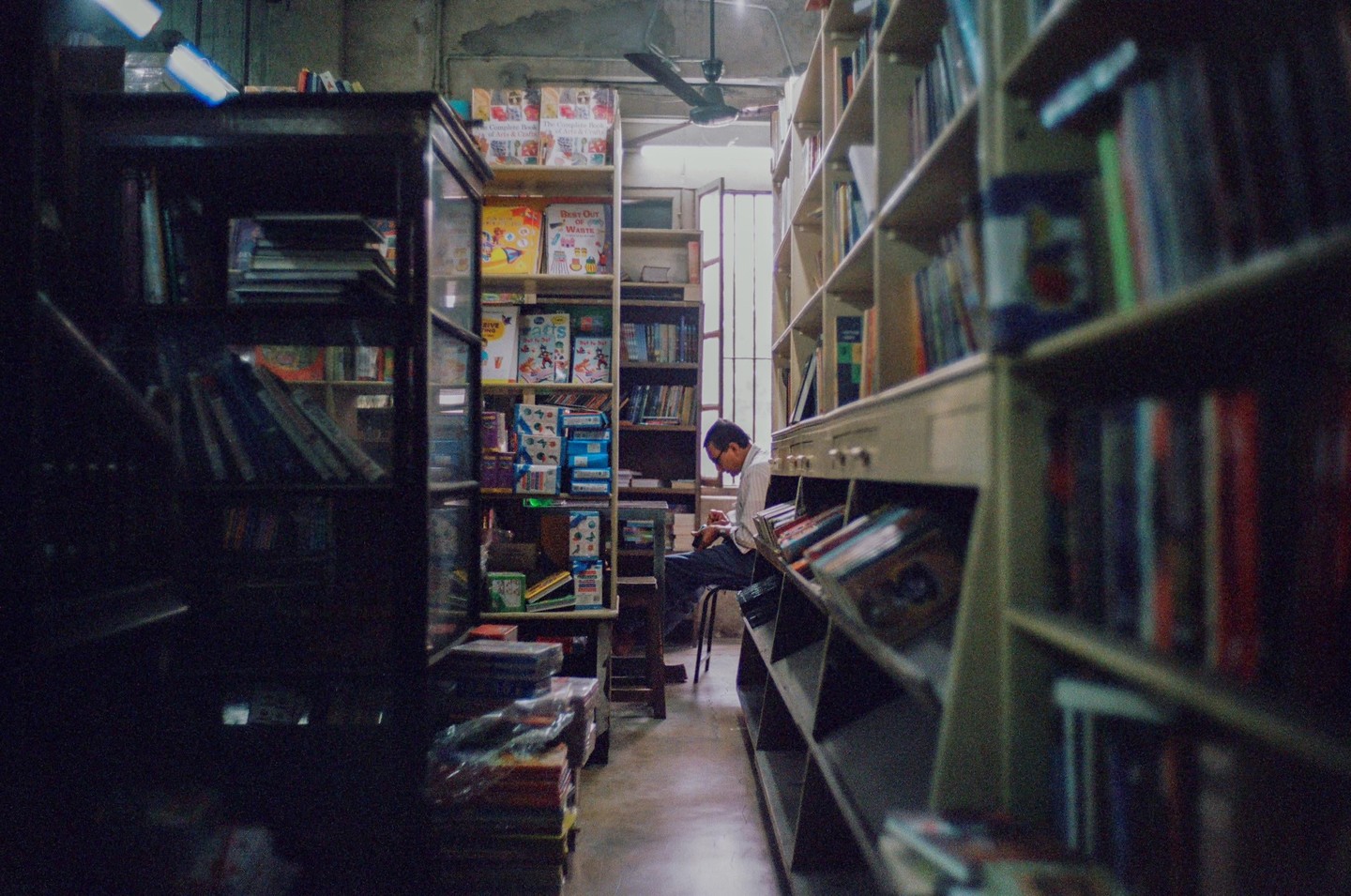 A man sits on a stool in a narrow aisle between tall bookshelves in a bookstore.