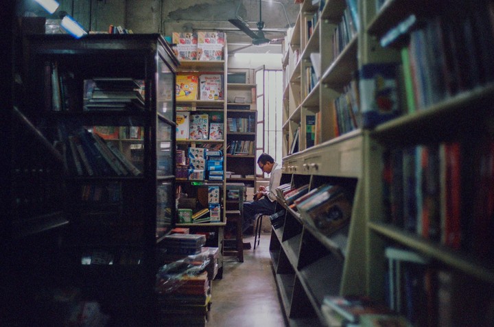 A man sits on a stool in a narrow aisle between tall bookshelves in a bookstore.