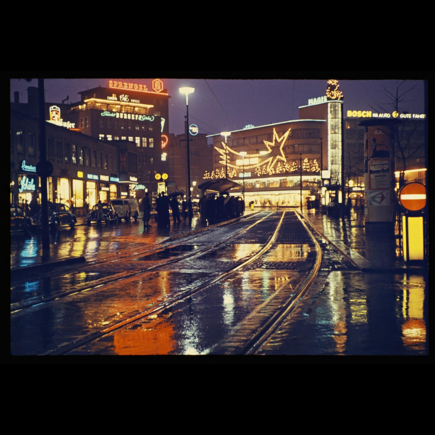 A street with tram tracks is wet from rain, reflecting the neon lights of buildings at night.