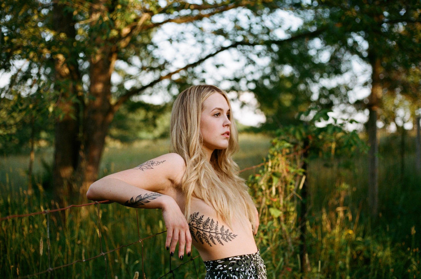A woman with long blonde hair and tattoos on her arm and torso leans on a wire fence in a field.