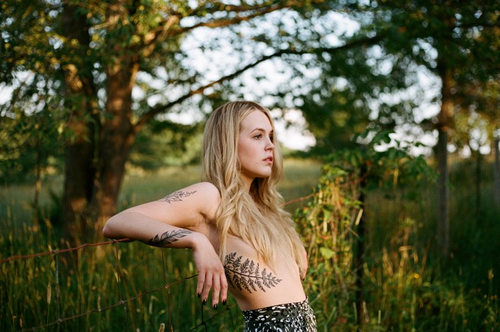 A woman with long blonde hair and tattoos on her arm and torso leans on a wire fence in a field.