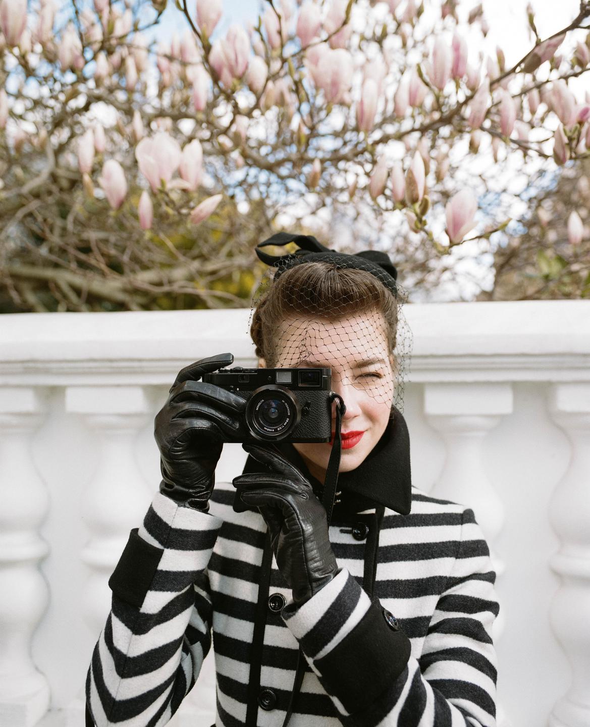 A woman in a striped coat and veil hat holds a camera to her eye, with magnolia blossoms in the background.