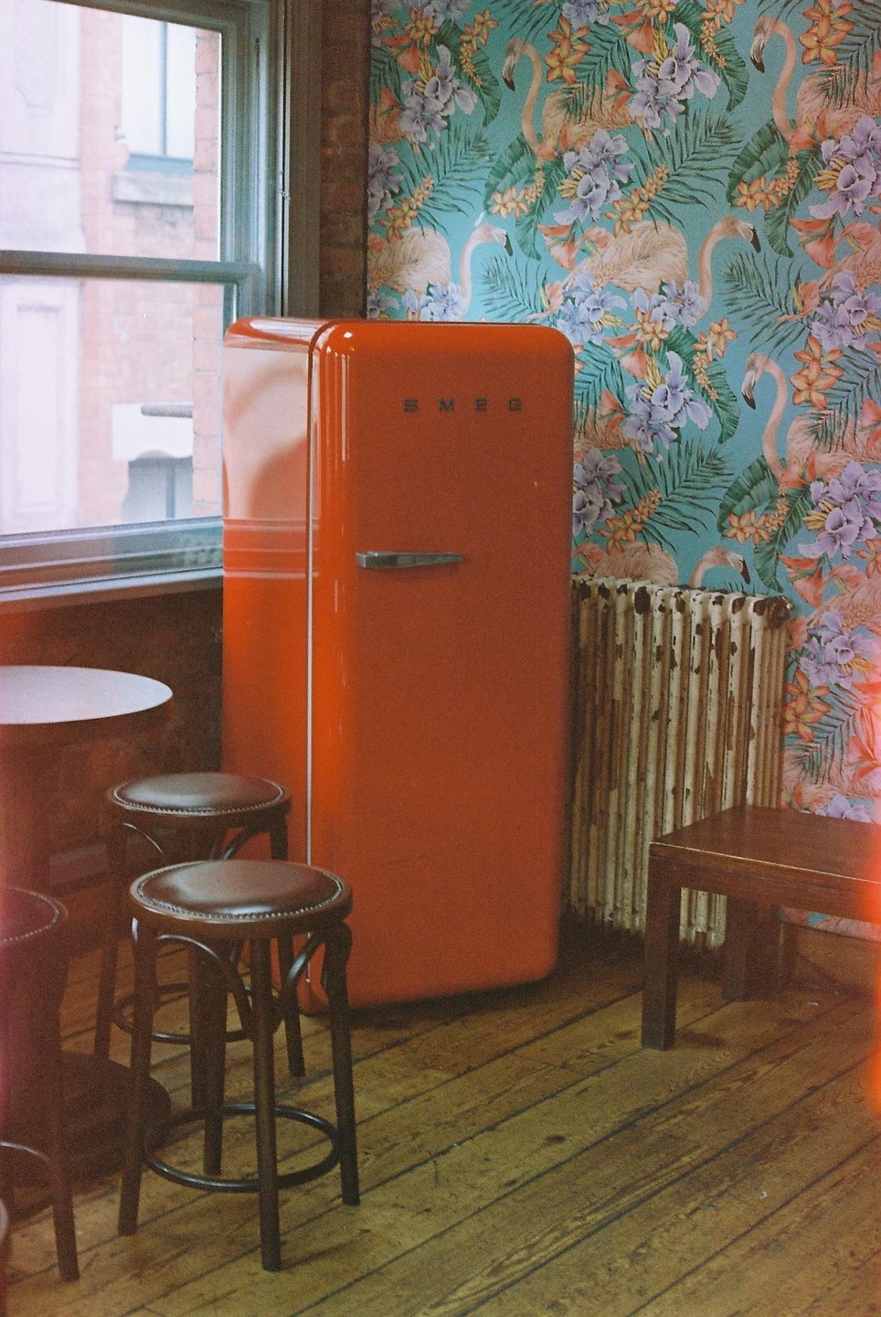 A red refrigerator stands in a cafe corner next to a window, with stools and a table in the foreground and flamingo wallpaper on the wall.