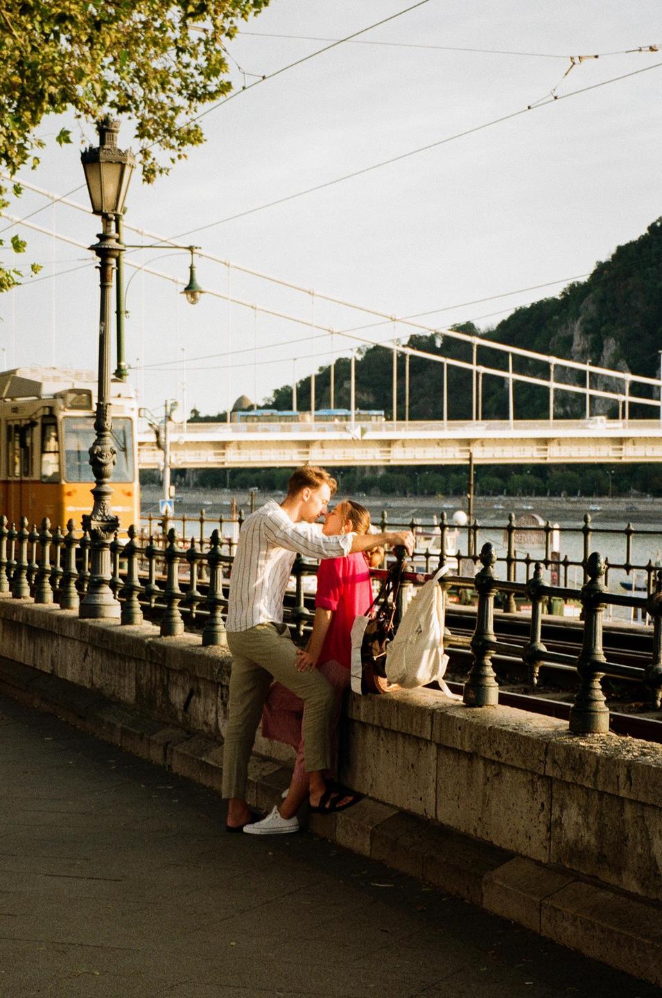 A couple kisses by a stone wall next to tram tracks, with a bridge and a mountain in the background.