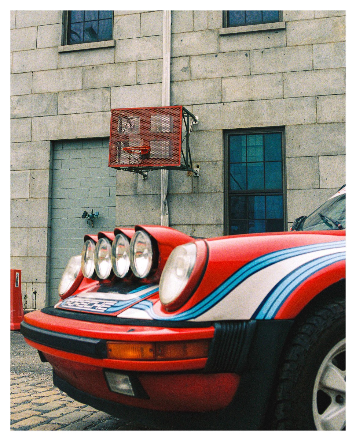 A red car with racing stripes and multiple rally lights is parked in front of a brick building with a basketball hoop.