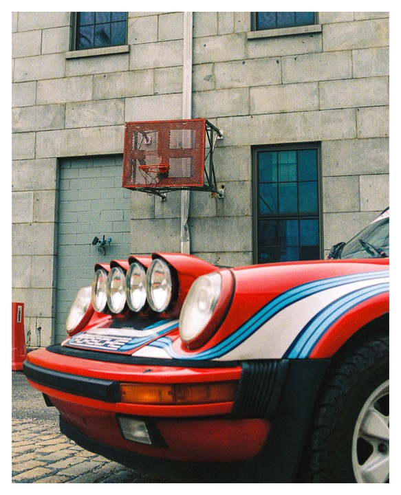 A red car with racing stripes and multiple rally lights is parked in front of a brick building with a basketball hoop.