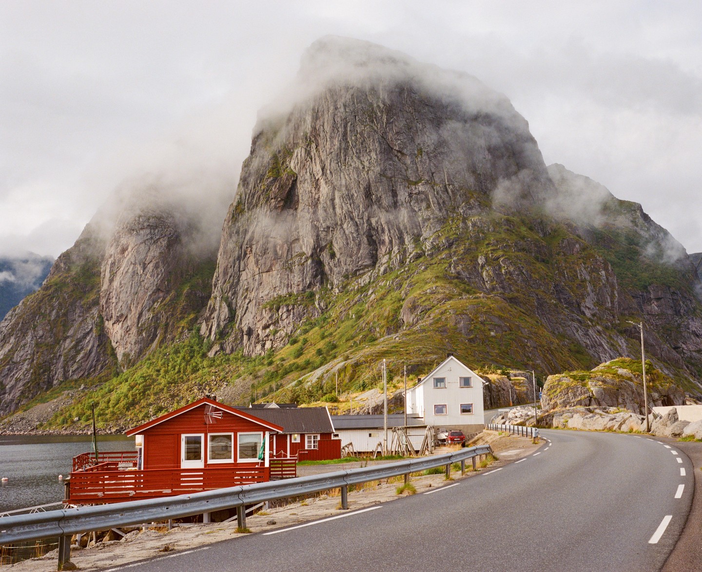 A winding road passes by red and white houses at the base of a large, fog-covered mountain.