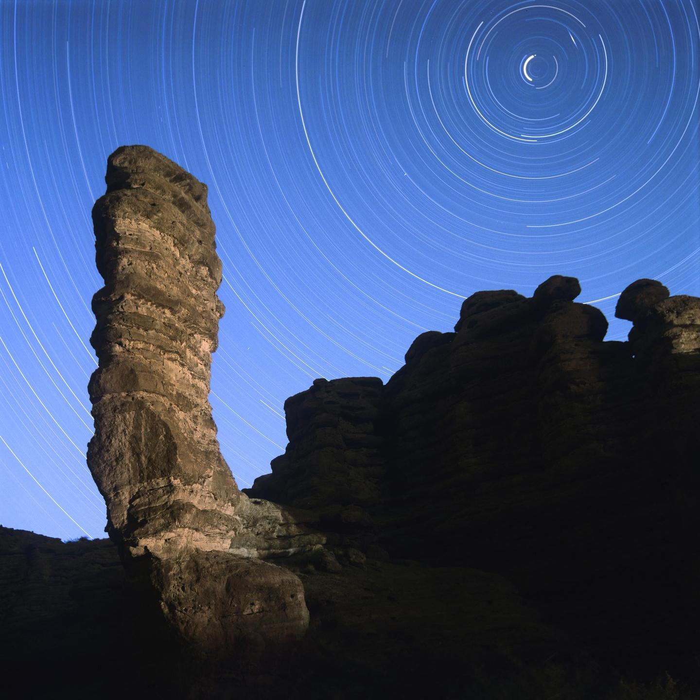 A tall rock formation stands against a night sky filled with star trails.