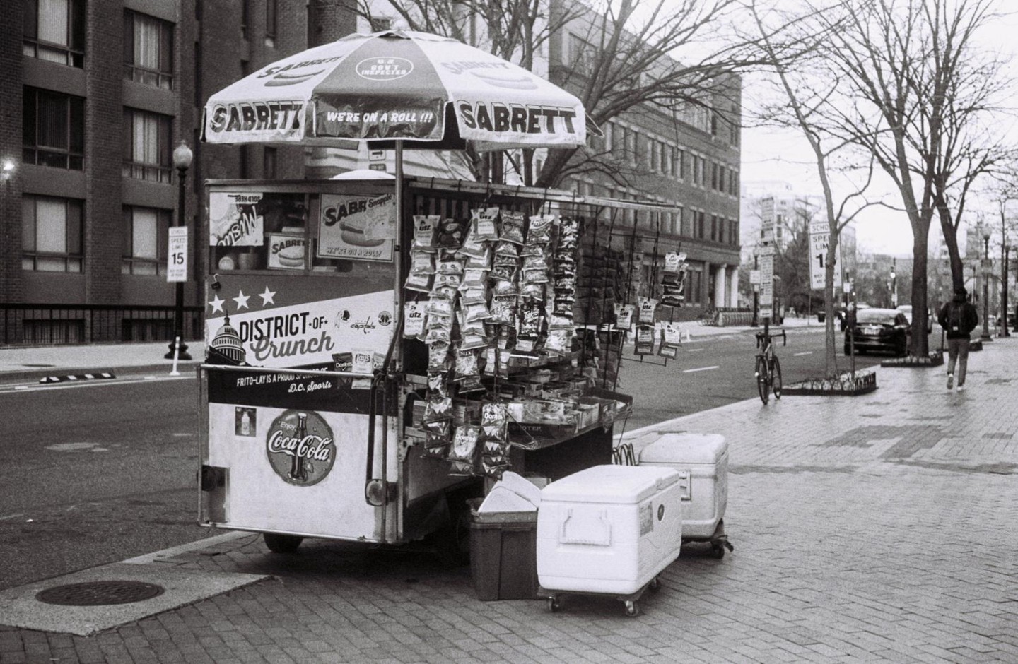 A food cart with an umbrella is parked on a city sidewalk next to a street with buildings and trees.