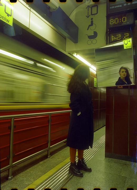 A woman stands on a subway platform as a train speeds by, with her reflection visible in a mirror.