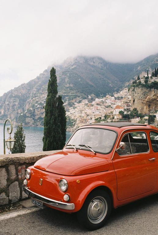 An orange car is parked on a road overlooking a coastal town and mountains.