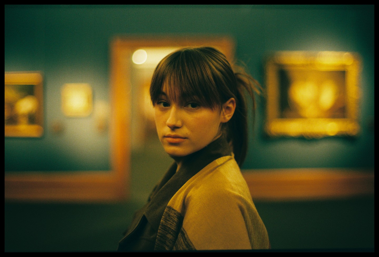 A young woman with bangs and a ponytail looks over her shoulder in a museum with blurred paintings in the background.