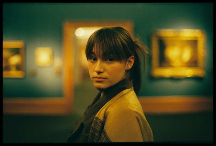 A young woman with bangs and a ponytail looks over her shoulder in a museum with blurred paintings in the background.
