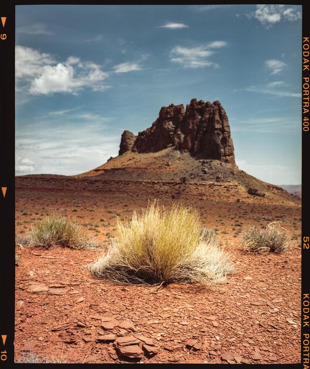 A large rock formation rises from a desert landscape with sparse vegetation under a blue sky with clouds.
