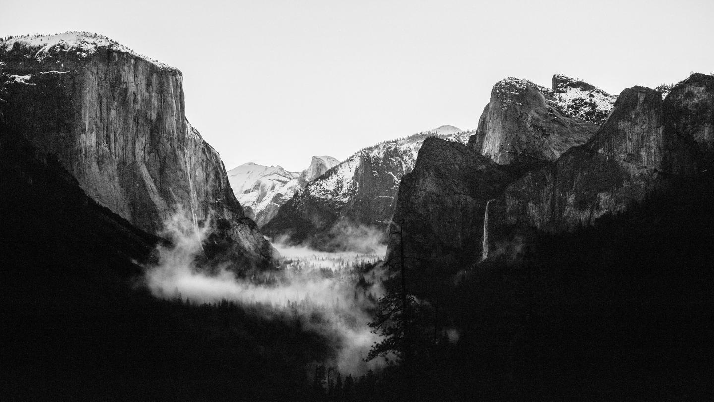 A black and white landscape shows a valley with fog and a waterfall surrounded by mountains.