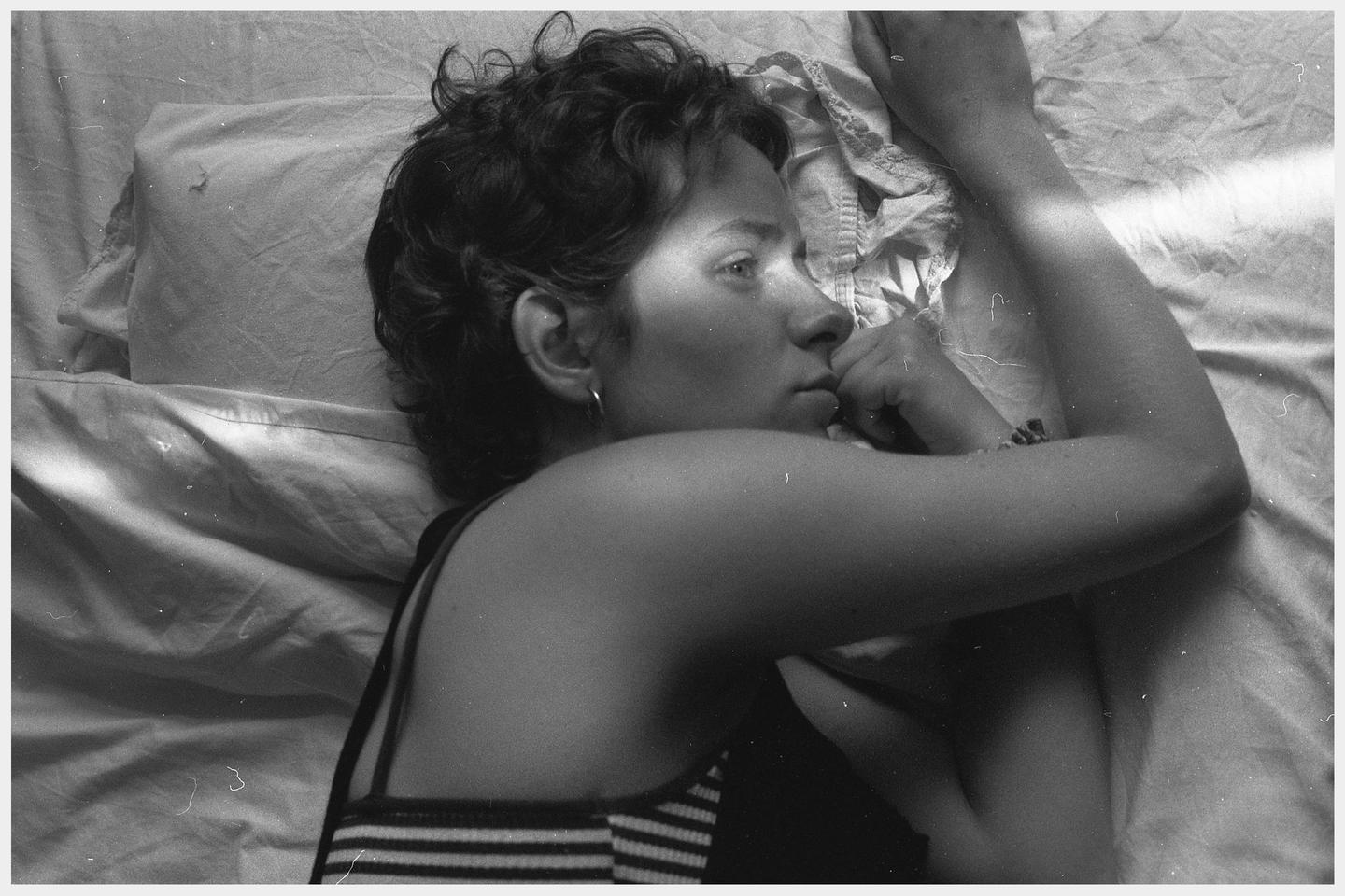 A woman with short curly hair lies on a bed, looking to the right with her hand near her mouth.