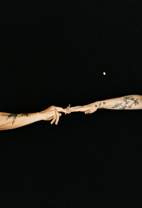Two tattooed arms reach towards each other, with one hand passing a lit cigarette to the other against a dark background with a small light source.