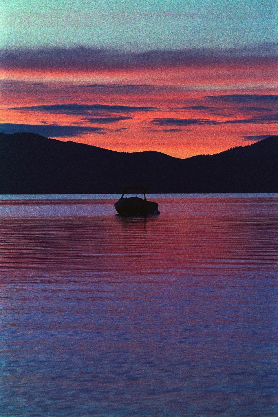 A boat floats on a lake with mountains in the background under a colorful sky at sunrise.