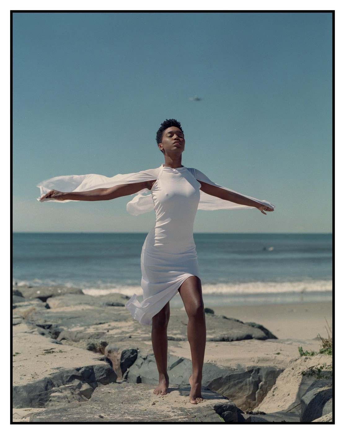 A woman in a white dress stands barefoot on rocks at the beach with her arms outstretched.