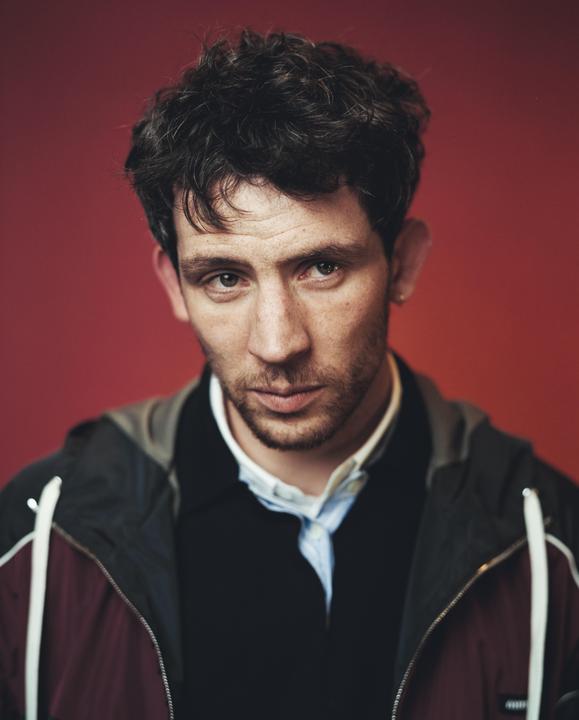 A man with dark, curly hair and a beard looks directly at the viewer against a red background.