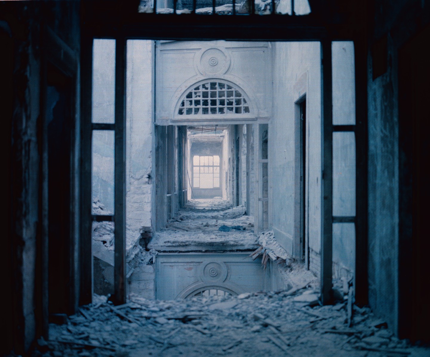 A long, narrow, and dilapidated hallway with debris on the floor leads to a bright window at the far end.