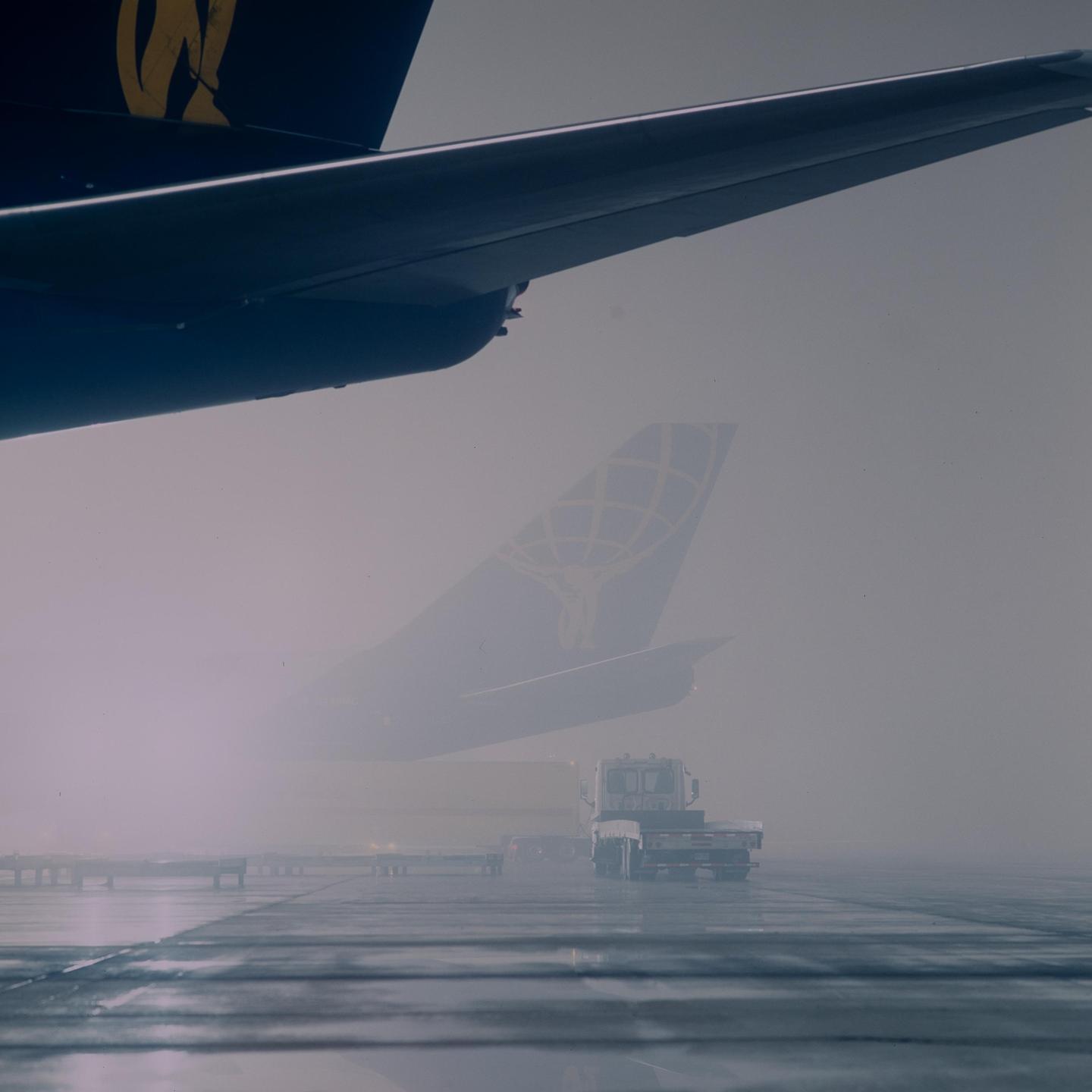 An airplane wing and tail are visible through a thick fog on an airport tarmac with a truck and trailer.
