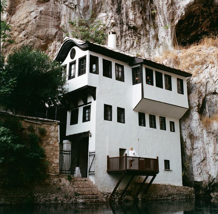 A white house with a dark roof and windows is built into a rocky cliff face above a body of water.