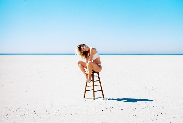 A woman in a white bikini sits on a stool on a vast white sand beach under a clear blue sky.